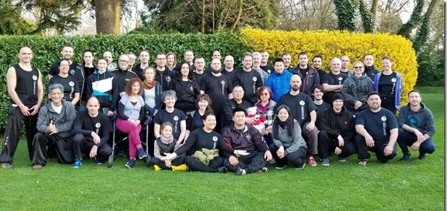 Group London 2019 Foto der Zhong Xin Dao Trainingsgruppe in London. 46 Menschen auf ca. 15 Ländern knien, sitzen und stehen auf einer Wiese vor einer Hecke.